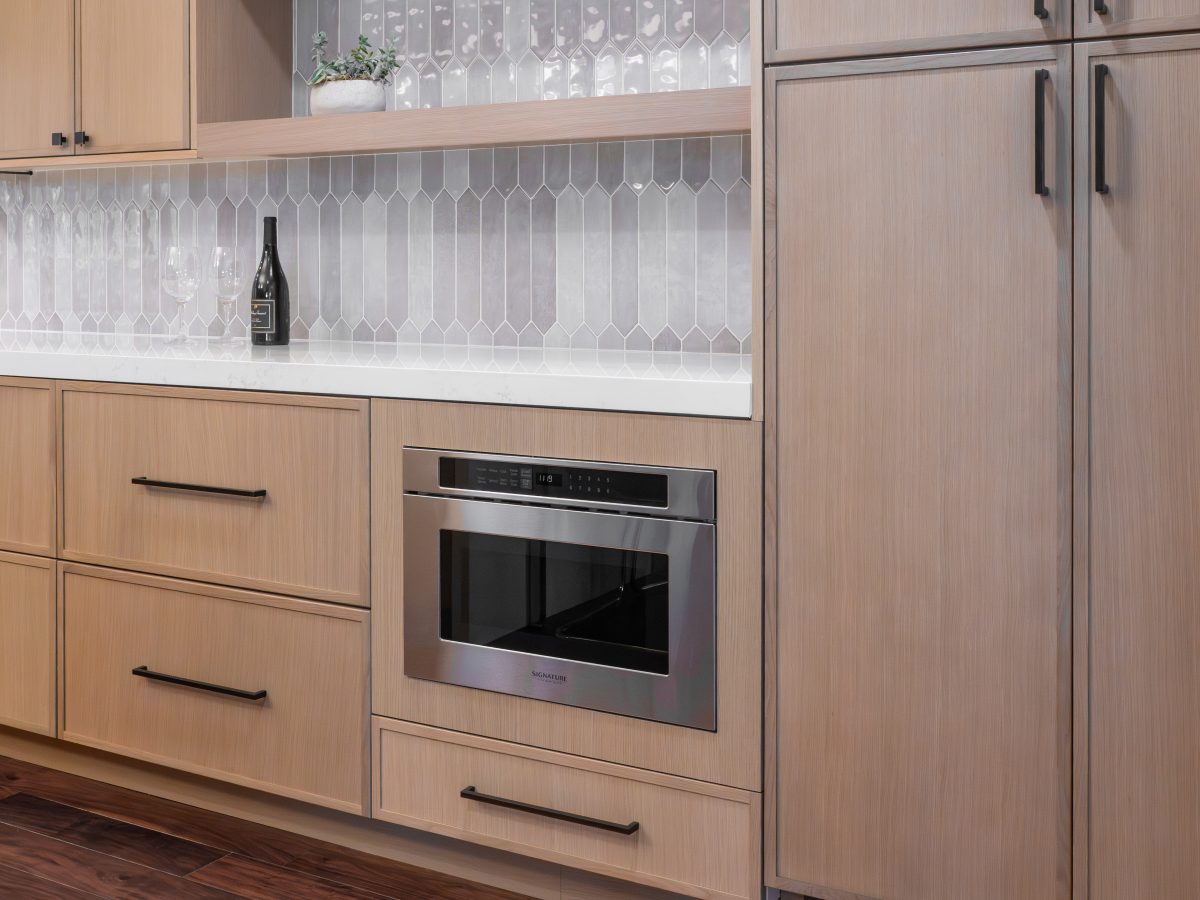 Kitchen with warm stained oak slim shaker cabinetry, dark bronze hardware, and picket-shaped backsplash tile — Reveal Interior Design Las Vegas