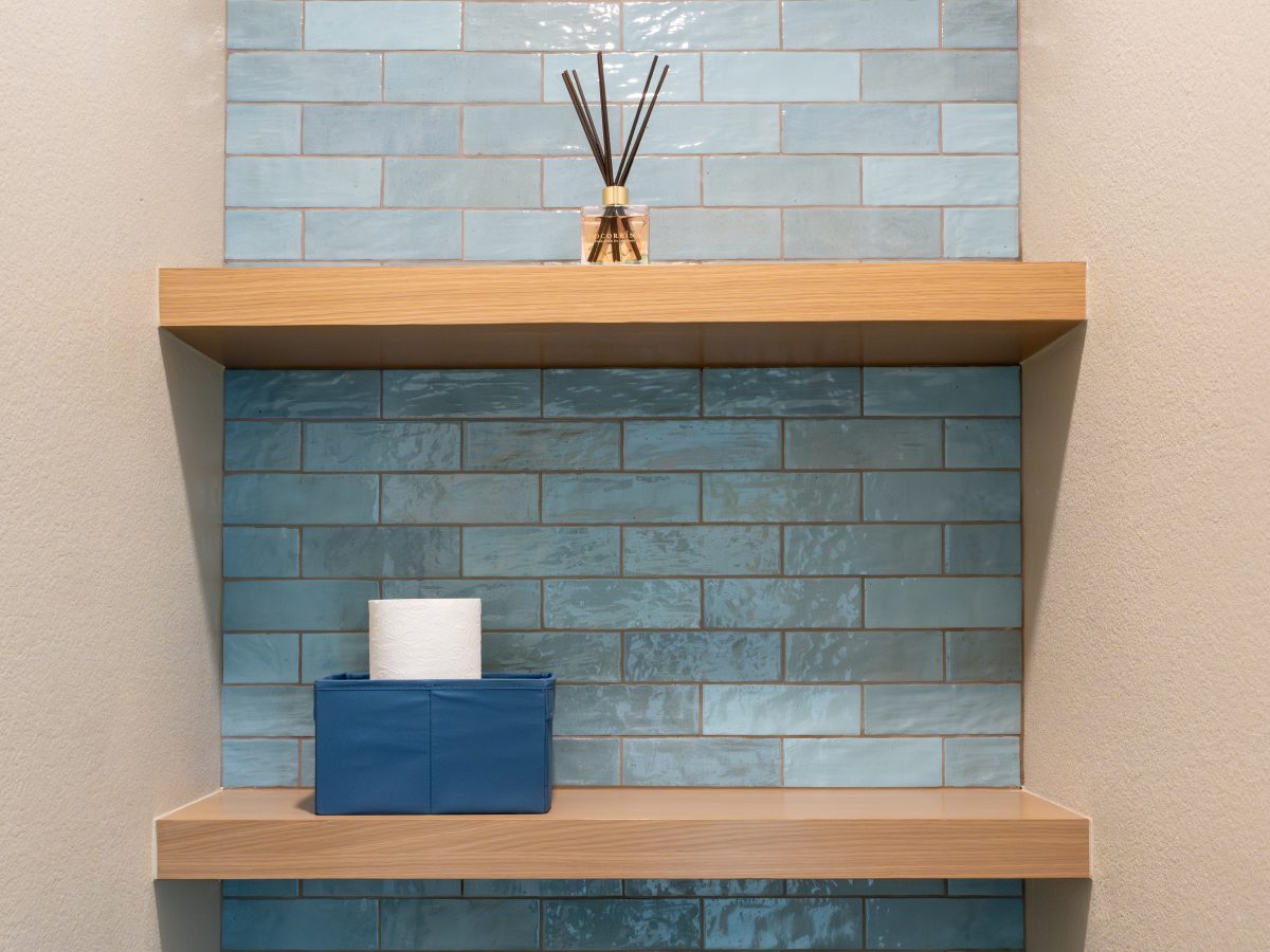 Toilet niche with handmade teal subway tile, warm oak floating shelves, and reed diffuser — Reveal Interior Design Las Vegas
