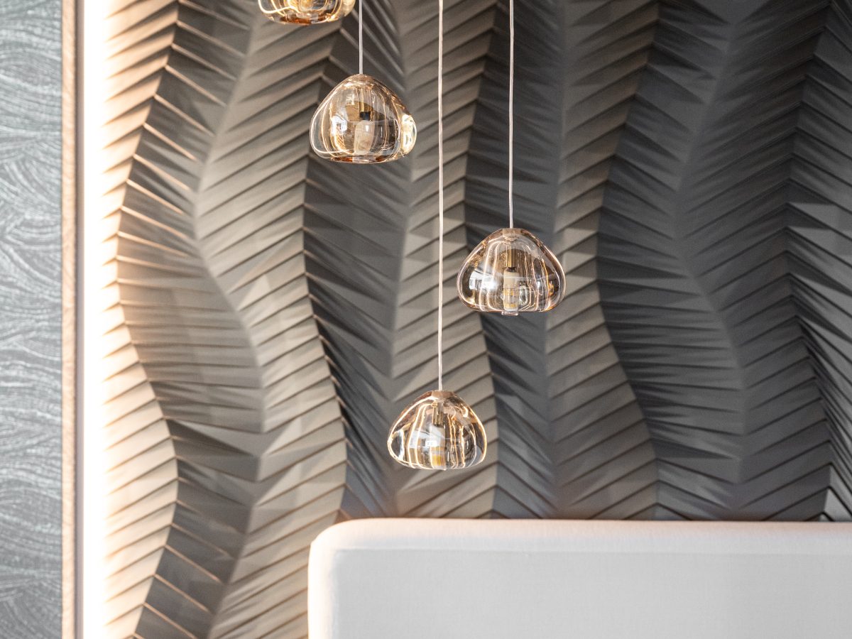 Primary bedroom with gold bronze textured wall panels, multi-drop pendants, and linear fireplace — Reveal Interior Design Las Vegas