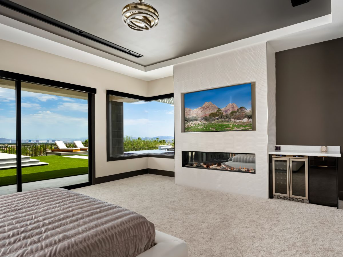 Primary bedroom with linear fireplace, recessed TV, corner glass walls and city views — Reveal Interior Design Las Vegas