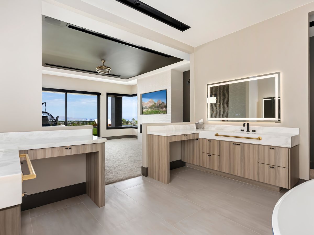 Primary bathroom with warm oak-look vanities, marble-look countertops, and LED mirrors opening to bedroom views — Reveal Interior Design Las Vegas