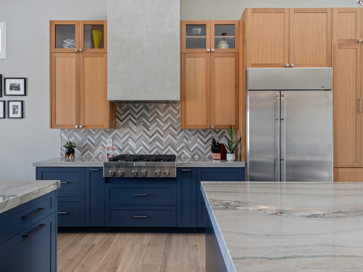 Kitchen with blue base cabinets, warm oak uppers, quartzite countertops, and herringbone mosaic backsplash — Reveal Interior Design Las Vegas