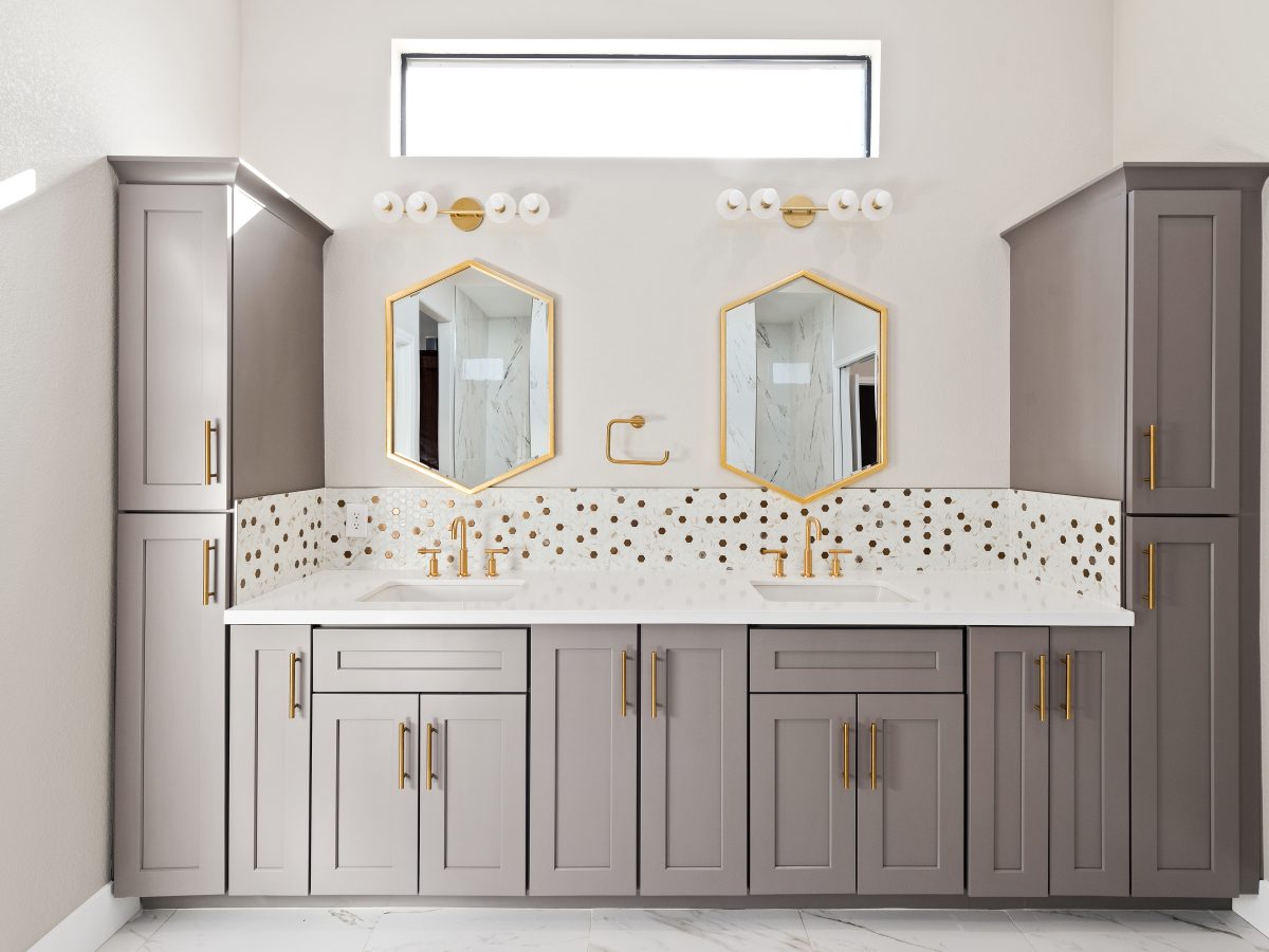 Primary bathroom double vanity with taupe shaker cabinets, gold fixtures, and geometric mirrors — Reveal Interior Design Las Vegas