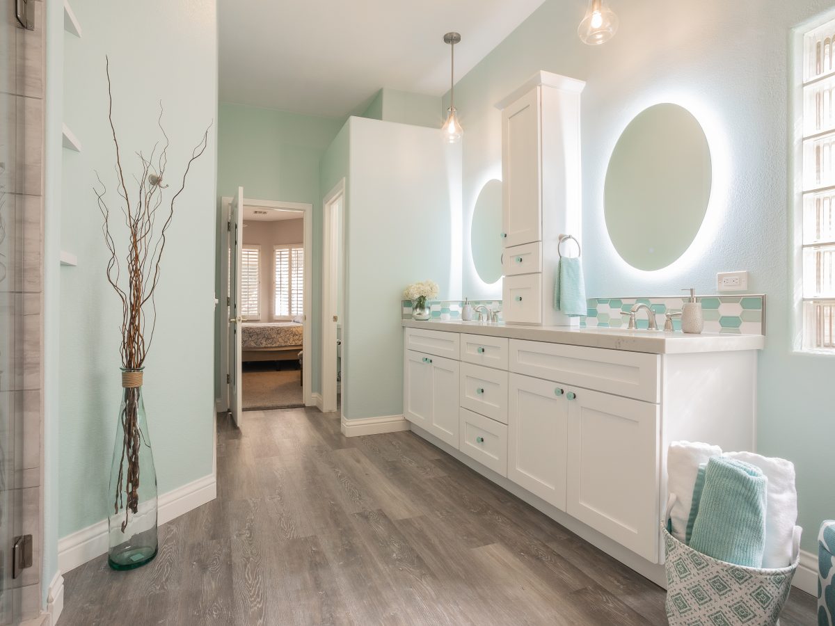 Coastal primary bathroom with backlit round mirrors, white quartz vanity, and teal hex mosaic tile — Reveal Interior Design Las Vegas