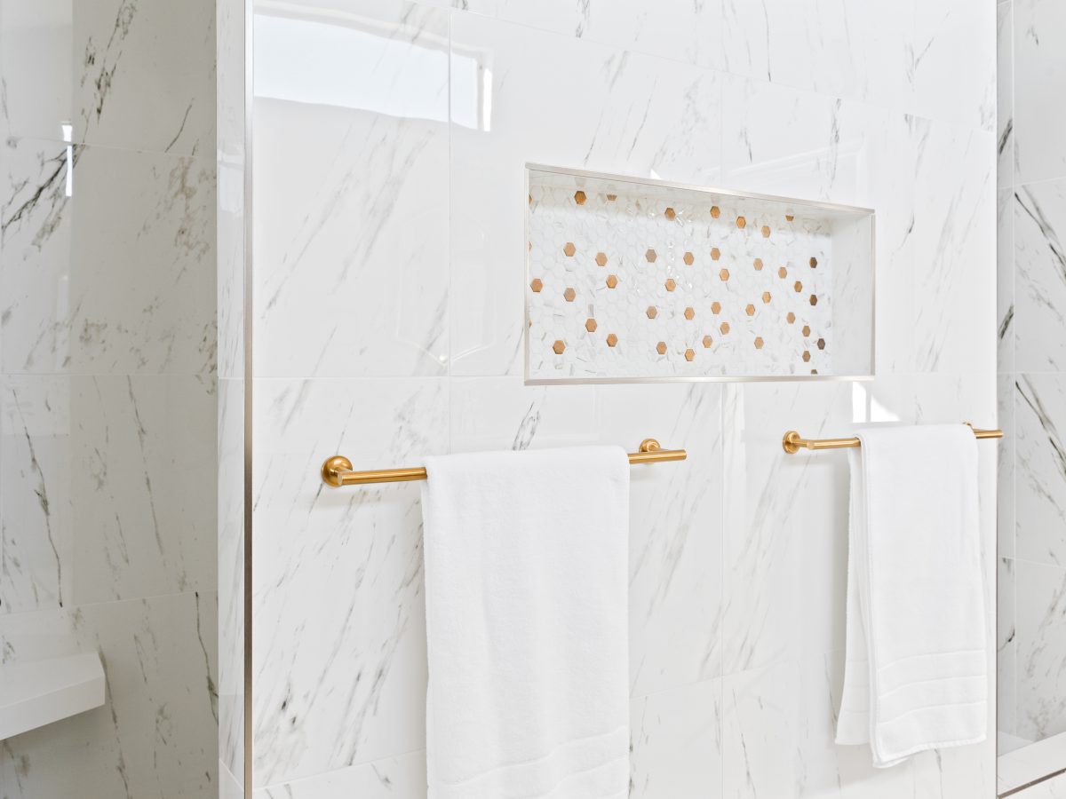 Primary bathroom shower wall with Calacatta-look porcelain tile, gold hexagon mosaic niche, and brushed gold towel bars — Reveal Interior Design Las Vegas