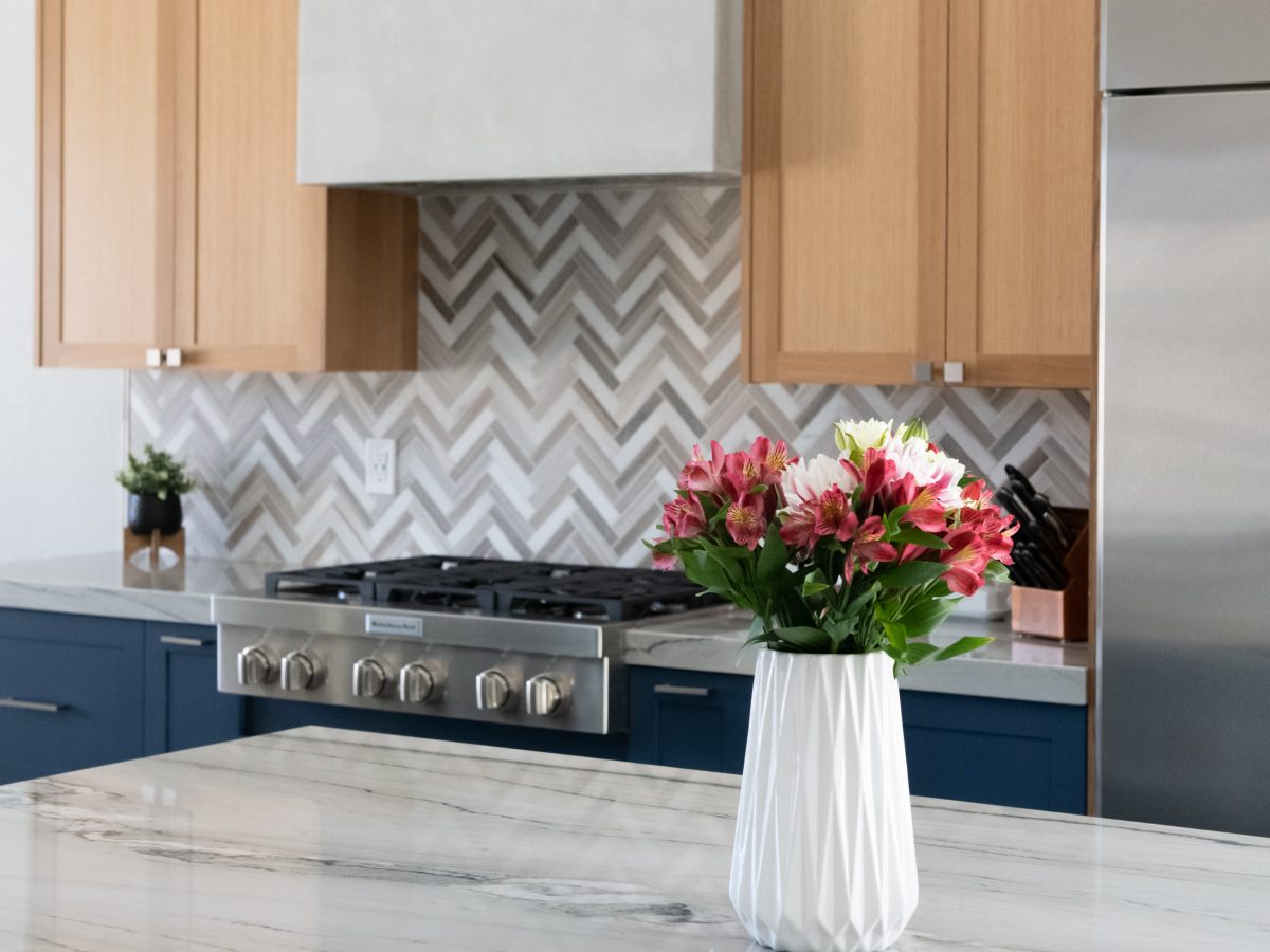 Kitchen with dark blue base cabinets, warm oak uppers, quartzite island countertop, and herringbone mosaic backsplash — Reveal Interior Design Las Vegas