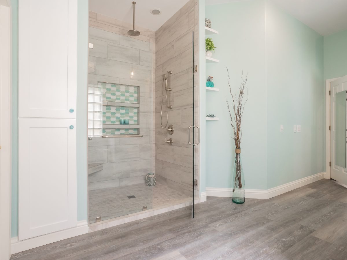Beach-inspired primary bathroom with frameless glass shower, stone-look porcelain tile, and wood-look plank flooring — Reveal Interior Design Las Vegas