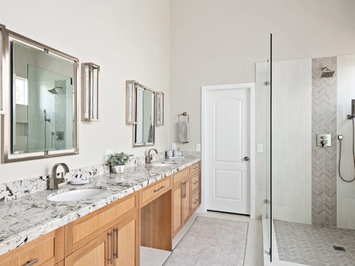 Primary bathroom with warm oak vanity, granite countertops, and glass-enclosed shower — Reveal Interior Design Las Vegas